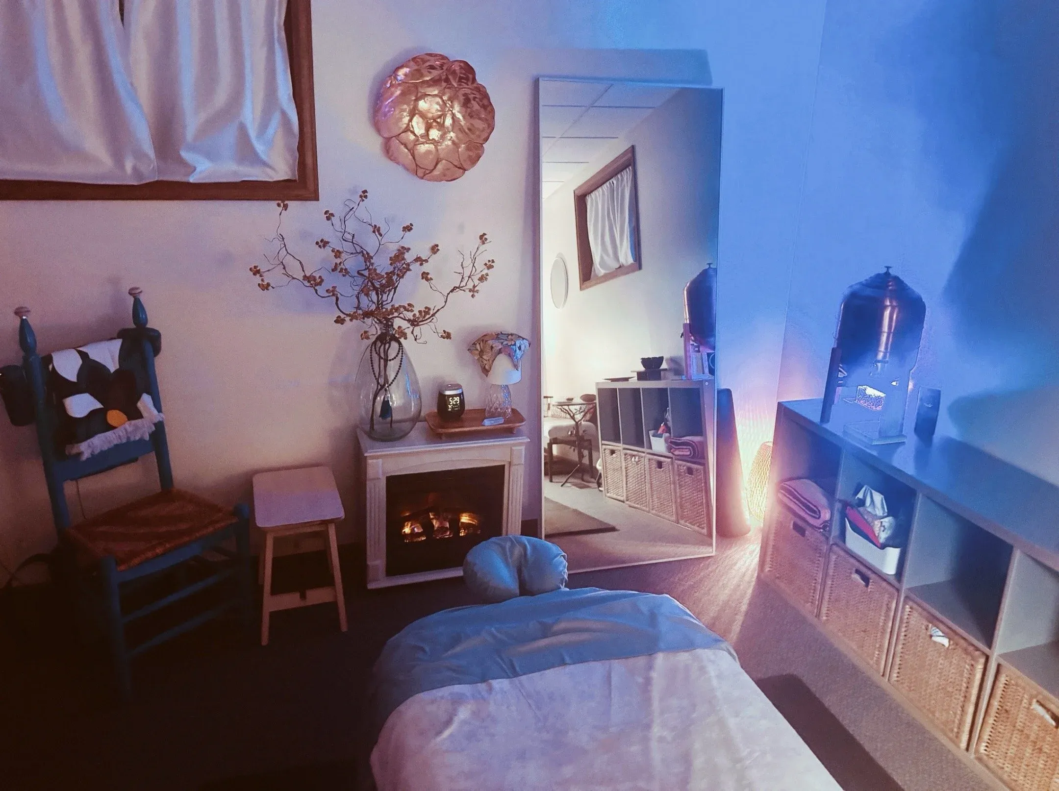 Treatment room at warm dusk lighting, massage table with crisp linens, fireplace, blue rustic chair, dried branches